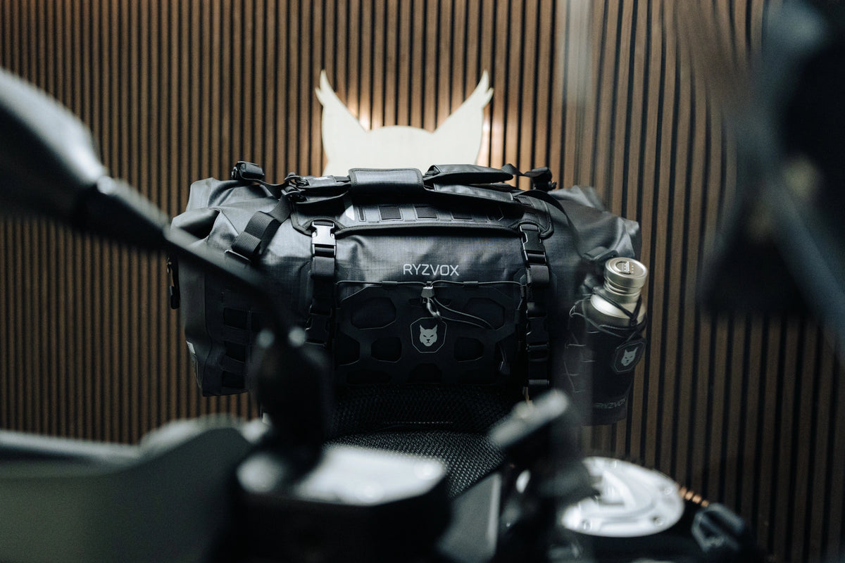 RYZVOX FAR Duffel Bag 40L strapped to the tail of a motorcycle with a bottle holder attached, RYZVOX lynx logo on the wall behind