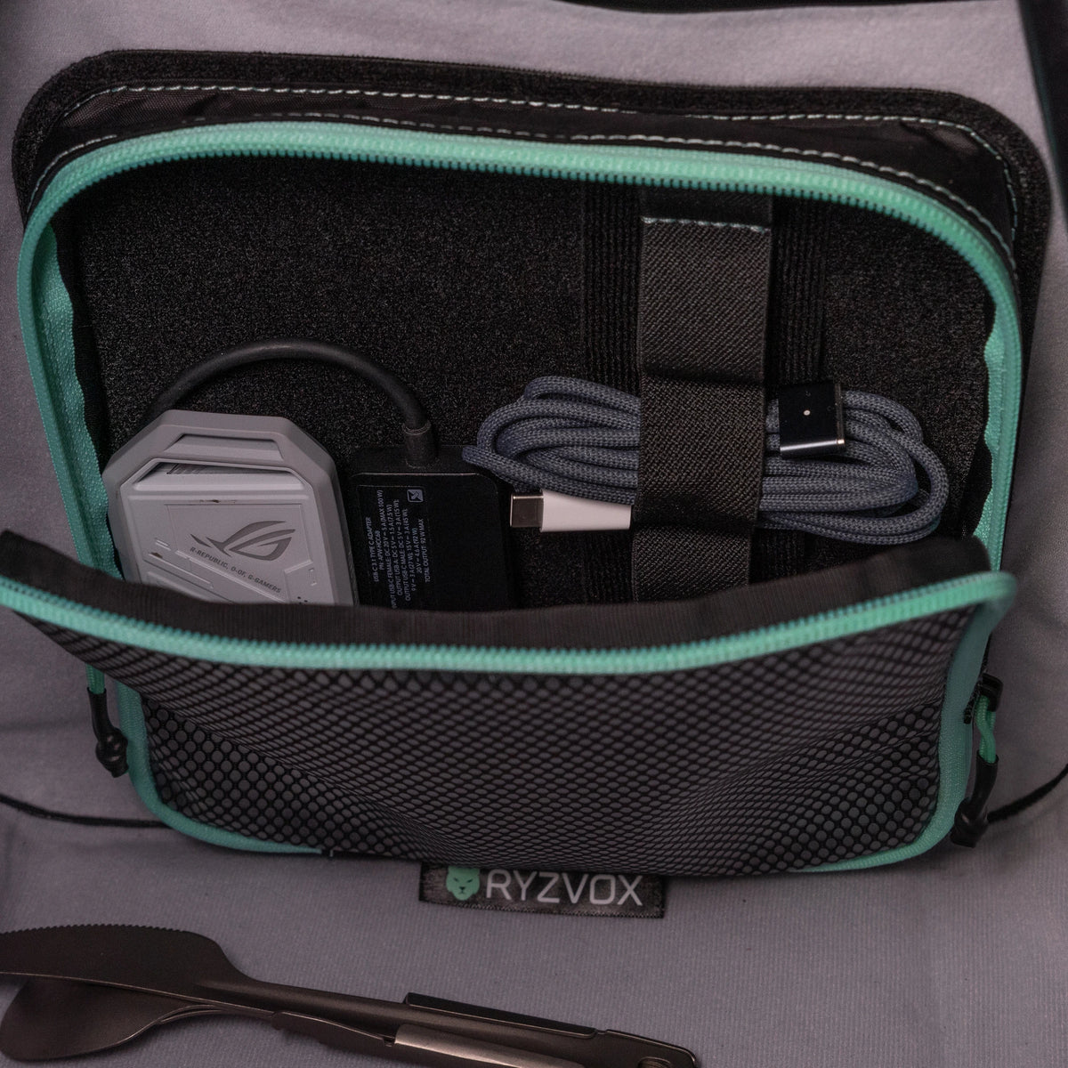 Mesh pouch open inside a RYZVOX bag, holding a laptop charger and cables